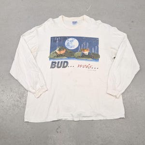 Puede incluir: Camiseta de manga larga color crema con un gráfico de dos ranas sobre nenúfares bajo una luna llena. La camiseta tiene el texto "BUD... weis..." impreso debajo.