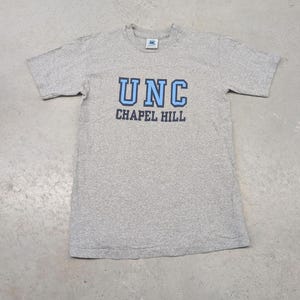 Könnte beinhalten: Graues T-Shirt mit dem Text "UNC CHAPEL HILL" in blauen Blockbuchstaben. Das Shirt hat einen Rundhalsausschnitt und kurze Ärmel. Das T-Shirt besteht aus einem weichen, bequemen Material.