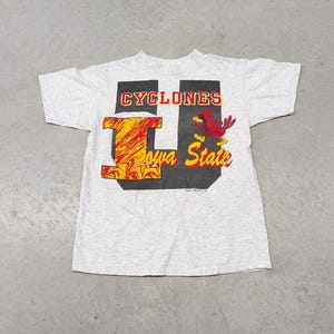 Può includere: Maglietta grigia con una grafica rossa e gialla della lettera "I" e un cardinale rosso con il testo "CYCLONES Iowa State".