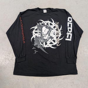 May include: A black long-sleeve t-shirt featuring a white circular graphic with a central figure and text. The right sleeve displays the red text "LA TRAVAGANZA" and the left sleeve features the white text "REBEL" in a bold, blocky font style.