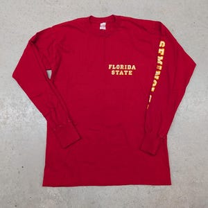 May include: A long-sleeved red shirt with the words "FLORIDA STATE" in gold lettering across the chest. The right sleeve has the word "SEMINOLES" printed vertically in gold. The shirt is made of a soft material.