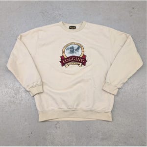 Pode incluir: Camisola creme com gola redonda e mangas compridas. A frente apresenta um emblema circular com as palavras "Vintage Canada Logging Circa 1900" e uma cena de exploração madeireira puxada por cavalos. Punhos e bainha canelados.
