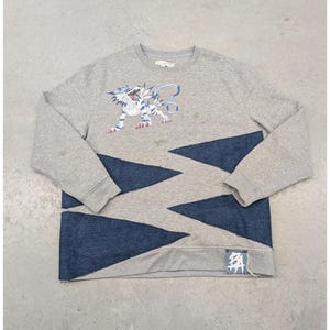 May include: A grey sweatshirt featuring a cartoon tiger in blue and white. The lower section has a zigzag pattern in grey and blue denim. A small tag with Japanese characters is attached to the hem.