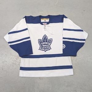May include: White and blue Toronto Maple Leafs hockey jersey. The jersey features a large team logo on the front, with blue stripes on the sleeves and a blue yoke. The jersey has a lace-up neckline.