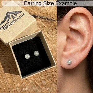 May include: A pair of small, round, glittery stud earrings displayed in a small, open cardboard box with a black interior. The box is labeled "Upstate Resin Works LLC." The earrings are also shown in a person's ear. The text "Earring Size Example" is at the top.