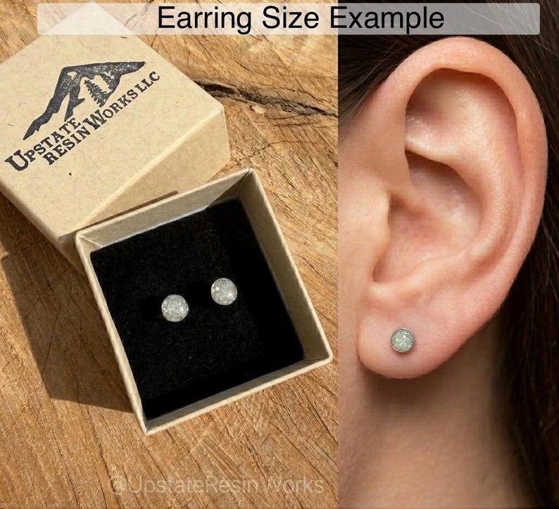 May include: A pair of small, round, glittery stud earrings displayed in a small, open cardboard box with a black interior. The box is labeled "Upstate Resin Works LLC." The earrings are also shown in a person's ear. The text "Earring Size Example" is at the top.