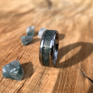 Op de afbeelding: Een zilveren ring met een groene en zilveren inleg. De ring ligt op een houten oppervlak met kleine groene stenen.