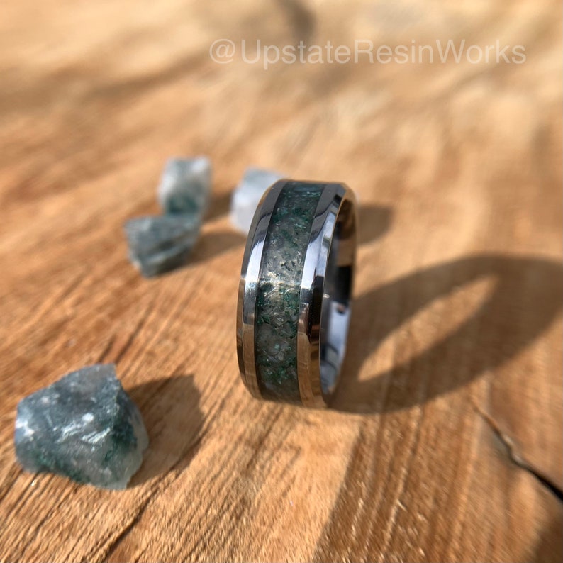 Op de afbeelding: Een zilveren ring met een groene en zilveren inleg. De ring ligt op een houten oppervlak met kleine groene stenen.