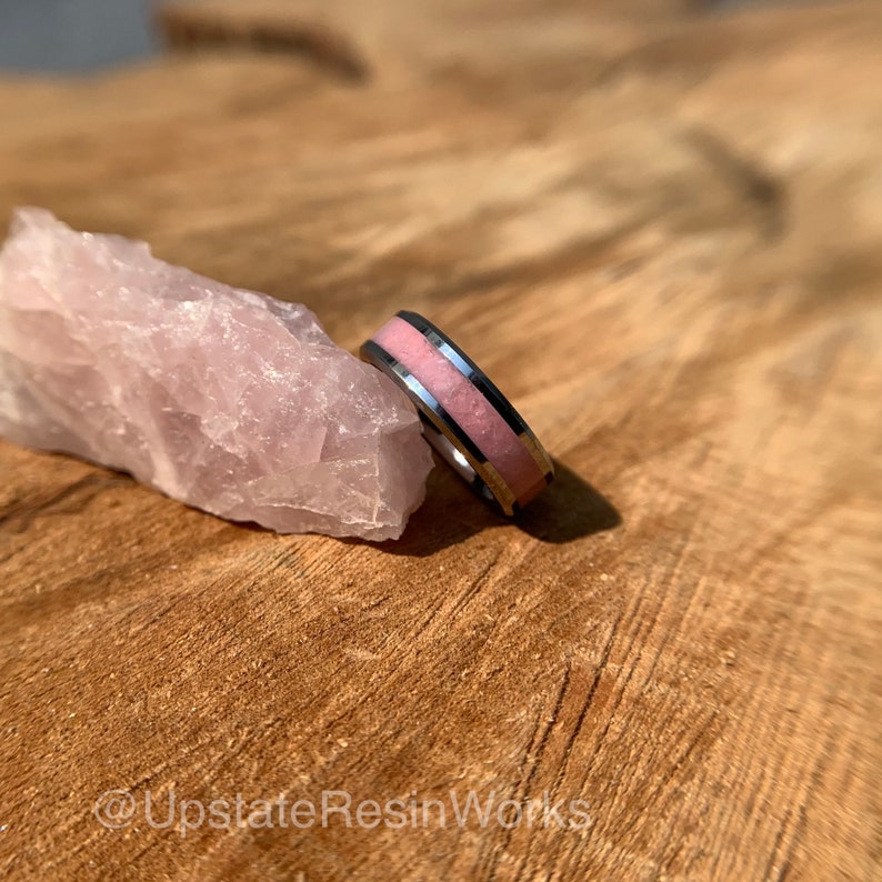 May include: A silver ring with a pink inlay. The ring is resting on a piece of rose quartz.