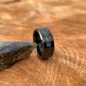 May include: Black tungsten carbide ring with a green and black inlay. The ring is on a wooden surface.