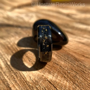 May include: A black tungsten carbide ring with a gold inlay. The ring has a brushed finish and is set against a wooden background.