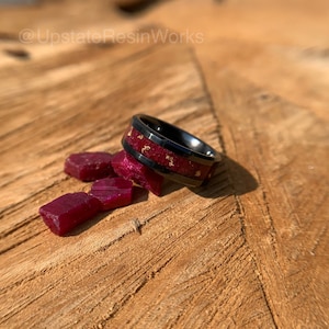 May include: A black ceramic ring with a red and gold inlay. The ring is sitting on a wooden surface with small red gemstones scattered around it.