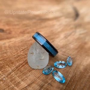 May include: A black ring with a blue inlay, resting on a white rock.  The ring is surrounded by several blue gemstones.
