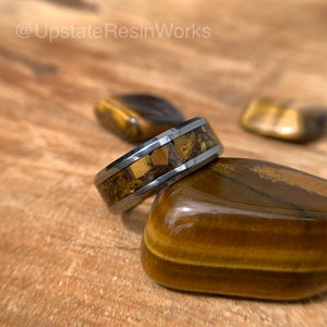 May include: A tungsten carbide ring with a band of embedded tiger's eye stones. The ring is resting on a tiger's eye stone.