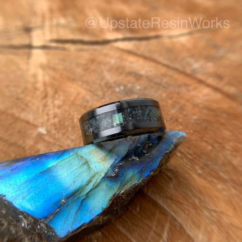 May include: A black ring with a band of iridescent green and black inlay. The ring is sitting on a blue and green rock.
