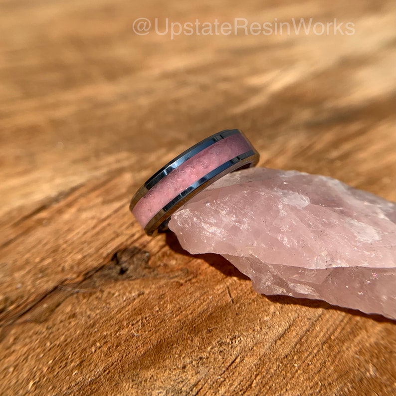 May include: A black tungsten ring with a pink resin inlay. The ring is sitting on a piece of rose quartz.