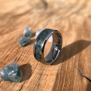 Op de afbeelding: Een ring van wolframcarbide met een groene en zilveren inleg. De ring ligt op een houten oppervlak met groene en witte stenen.
