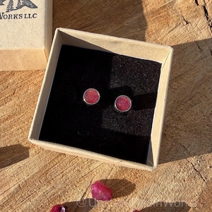 May include: A pair of round, pink gemstone stud earrings in a silver setting, presented in a small, open cardboard box with a black interior. The box sits on a wooden surface, with loose gemstones scattered nearby. The earrings are a simple, elegant design.
