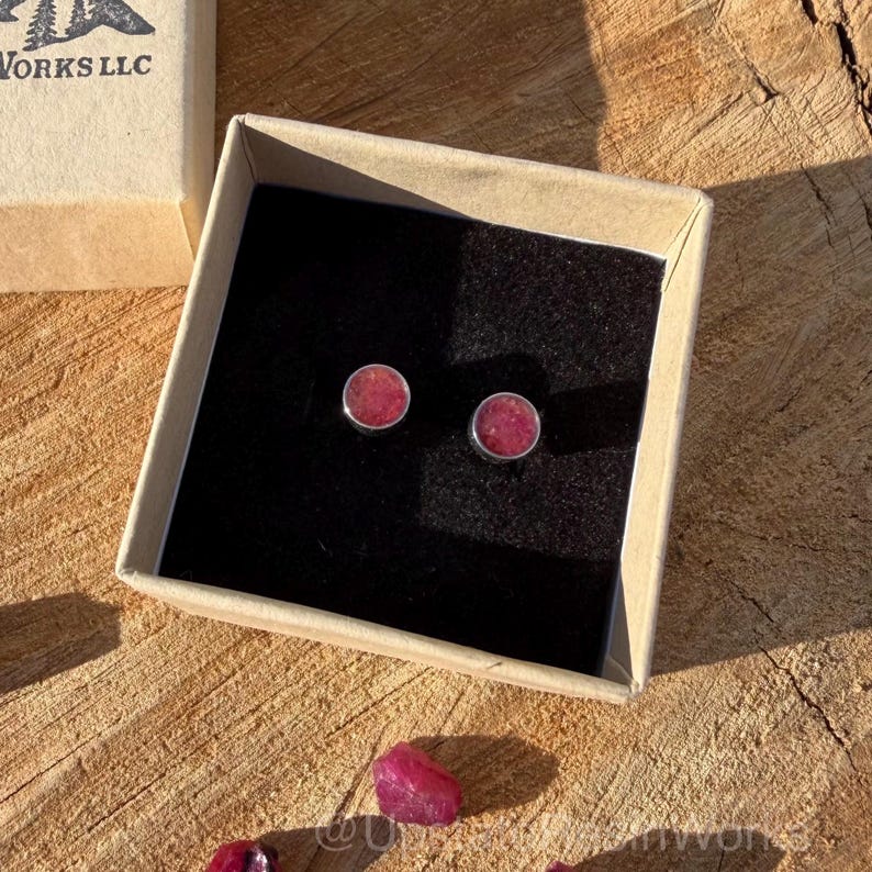 May include: A pair of round, pink gemstone stud earrings in a silver setting, presented in a small, open cardboard box with a black interior. The box sits on a wooden surface, with loose gemstones scattered nearby. The earrings are a simple, elegant design.