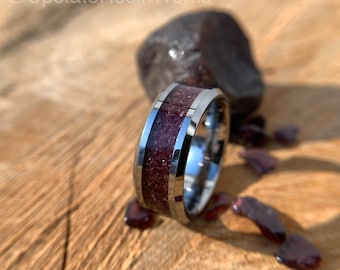 Puede incluir: Primer plano de un anillo de carburo de tungsteno con una incrustación de color rojo oscuro y plata. El anillo está sobre una superficie de madera con pequeños trozos de material rojo esparcidos a su alrededor.