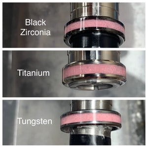 May include: Close-up of three metal rings with a pink band. The rings are labelled "Black Zirconia", "Titanium", and "Tungsten".