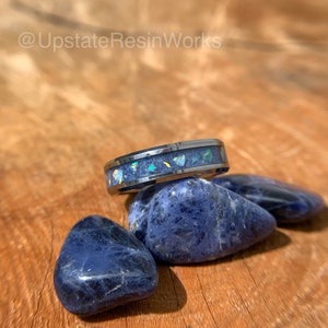 May include: A dark gray tungsten carbide ring with a blue and iridescent inlay. The ring is sitting on top of three blue stones.