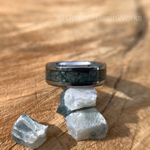 Op de afbeelding: Een zilveren ring met een groene en grijze inlay, rustend op een stapel groene en witte stenen. De ring ligt op een houten oppervlak. @UpstateResinWorks