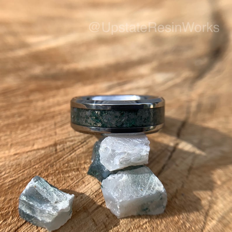 Op de afbeelding: Een zilveren ring met een groene en grijze inlay, rustend op een stapel groene en witte stenen. De ring ligt op een houten oppervlak. @UpstateResinWorks