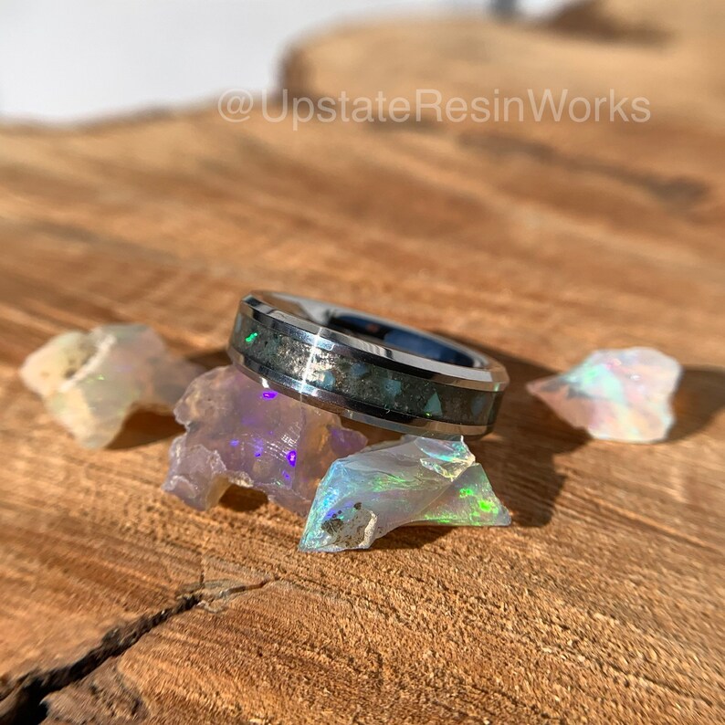May include: A silver ring with a black inlay and iridescent opal accents. The ring is sitting on a wooden surface with small pieces of opal.