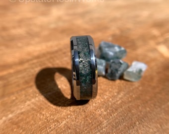 May include: A silver ring with a green and silver inlay. The ring is on a wooden surface with small green and white stones scattered around it.
