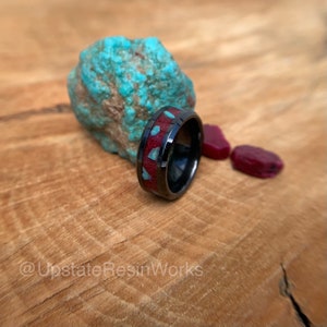 Puede incluir: Un anillo de tungsteno negro con una incrustación roja y turquesa. El anillo está sobre una superficie de madera con una roca turquesa y piedras rojas.