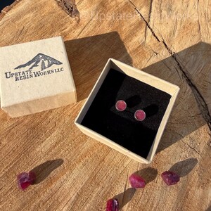 May include: A pair of small, round, pink stud earrings in a gift box with a black interior. The box is open, revealing the earrings. The box is sitting on a wooden surface. The box has the text "Upstate Resin Works LLC" and a mountain logo.