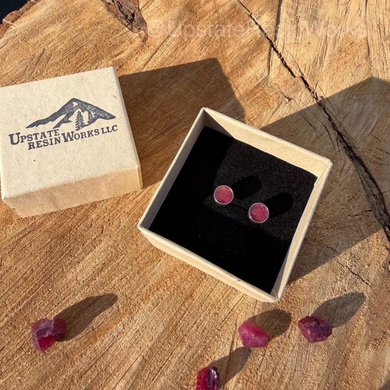 May include: A pair of small, round, pink stud earrings in a gift box with a black interior. The box is open, revealing the earrings. The box is sitting on a wooden surface. The box has the text "Upstate Resin Works LLC" and a mountain logo.