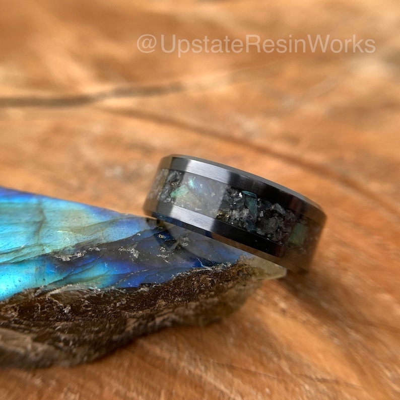 May include: A black tungsten carbide ring with a band of iridescent blue and green abalone shell and black glitter embedded in clear resin.