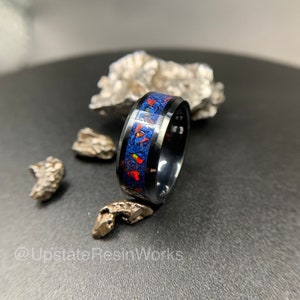 May include: A black ceramic ring with a blue and orange opal inlay. The ring is on a dark gray surface with small pieces of silver metal.