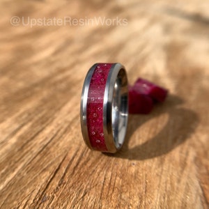 May include: A close-up of a dark red and silver ring with a textured band. The ring is made of titanium and resin.