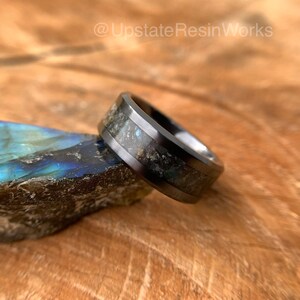 May include: A black tungsten carbide ring with a band of iridescent blue and black labradorite inlay. The ring is sitting on a piece of wood.