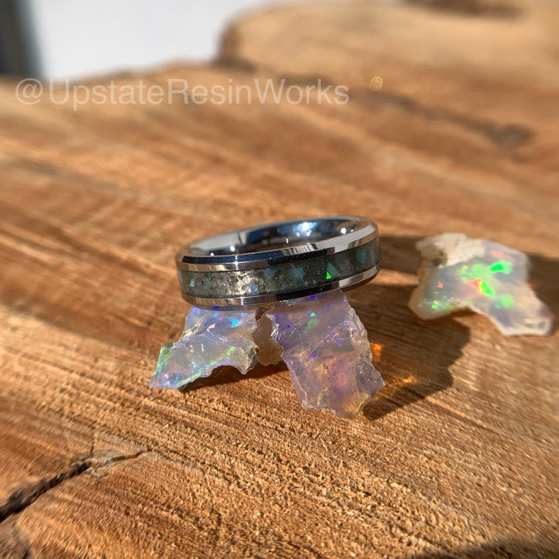 May include: A silver ring with a black and green inlay. The inlay is made of crushed opal and resin. The ring is sitting on a piece of opal.