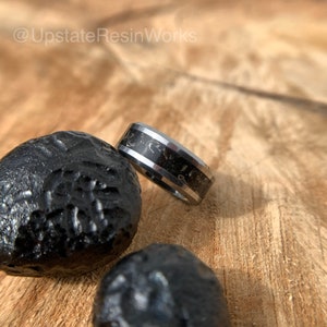 May include: A close-up of a ring with a black and silver design. The ring has a black center band with a marbled pattern, bordered by silver bands. The ring is next to three black, textured stones. The Etsy shop name "@UpstateResinWorks" is in the top left corner.