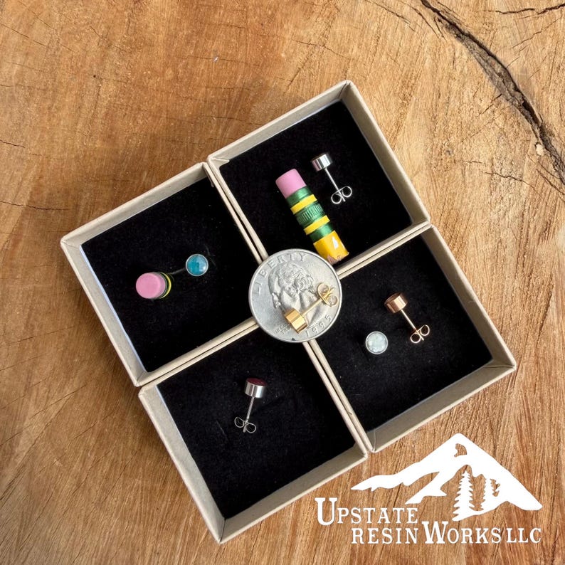 May include: Four small, square boxes with black interiors, each containing a pair of earrings. One box holds a pencil-shaped earring, another has a pink and blue earring, and two others have stud earrings. A quarter is in the center.