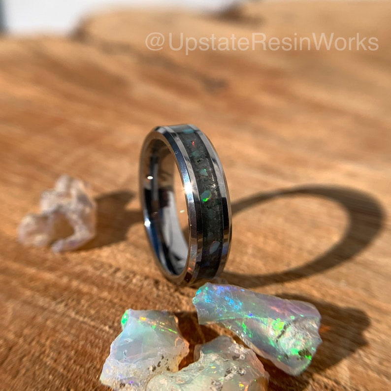 May include: A silver tungsten carbide ring with a black inlay and iridescent opal accents. The ring is on a wooden surface with pieces of opal.