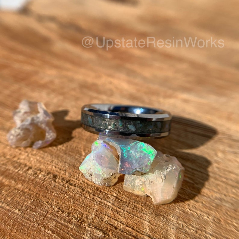 May include: A silver ring with a black band and a white inlay of iridescent opal. The ring is sitting on a pile of white opal stones.