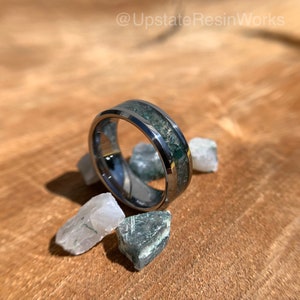 Op de afbeelding: Een zilveren ring van wolframcarbide met een groene en witte inlay. De ring is omgeven door kleine groene en witte stenen.
