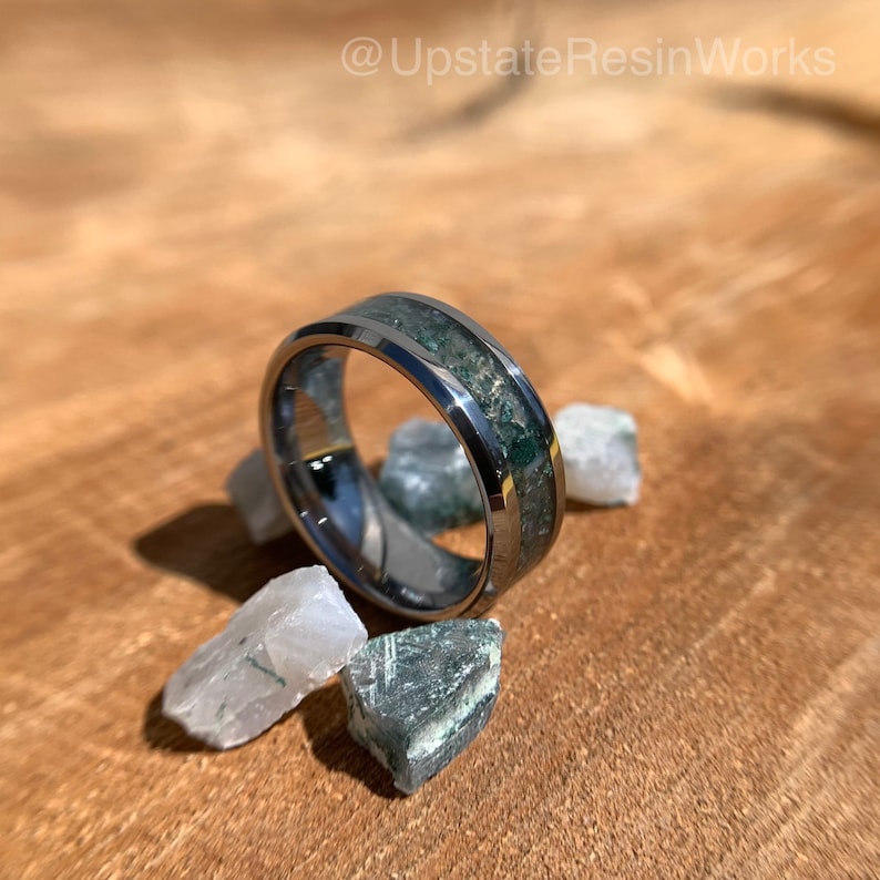 Op de afbeelding: Een zilveren ring van wolframcarbide met een groene en witte inlay. De ring is omgeven door kleine groene en witte stenen.