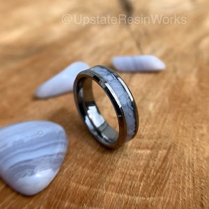 May include: A silver tungsten carbide ring with a white agate inlay. The ring is on a wooden surface with blue agate stones.