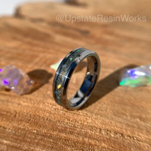 May include: A silver tungsten carbide ring with a black inlay and iridescent opal flakes. The ring is on a wooden surface.