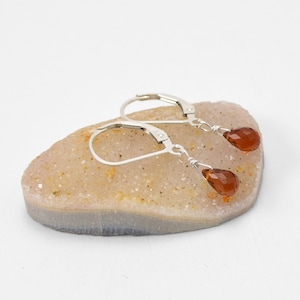 May include: Silver leverback earrings with teardrop-shaped, faceted, amber-colored gemstones. The earrings are displayed on a light-colored, textured stone against a white background. The gemstones are suspended from the silver leverback closures.