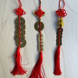 Könnte beinhalten: Drei rote und goldene chinesische Knotenschmuckstücke. Jedes Ornament hat eine rote Quaste, eine Reihe von Bronzemünzen und einen dekorativen Knoten. Die Ornamente sind an einer roten Schnur aufgehängt.