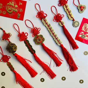 May include: A selection of red Chinese knot ornaments with gold coins and tassels. Several red envelopes with gold Chinese characters are also present. The ornaments vary in design, some featuring multiple coins strung together, and others with a single coin.