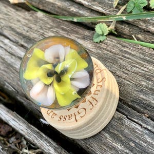 May include: A glass paperweight with a yellow and white flower design. The paperweight is sitting on a wooden base with the words "Glass Stuff" engraved on it.